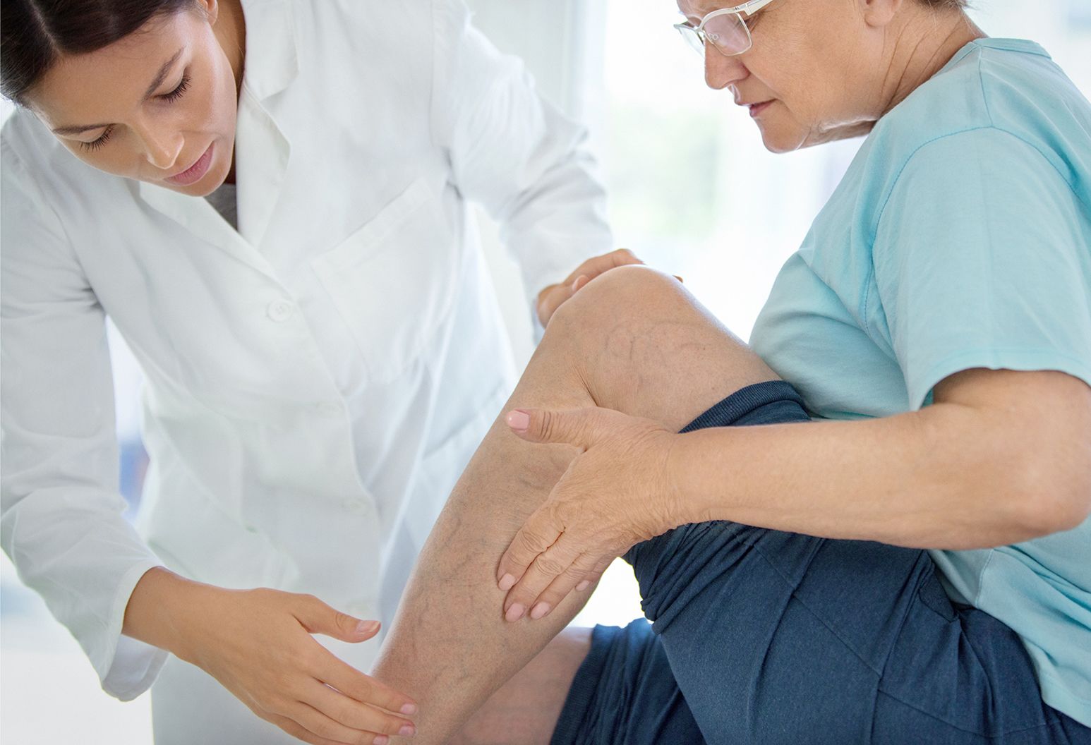 Closeup side view of female doctor massaging legs and calves of a senior female patient with visible varicose veins.