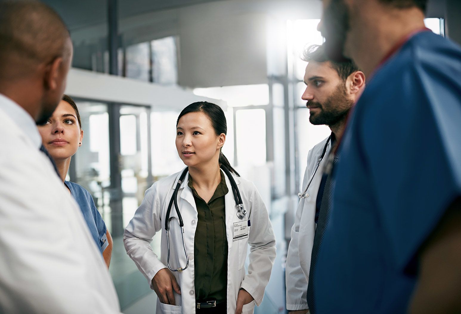 A team of doctors stand in a circle discussing a case. The focus is on a young asian female doctor wearing a dark shirt, white lab coat, and stethoscope around her neck.
