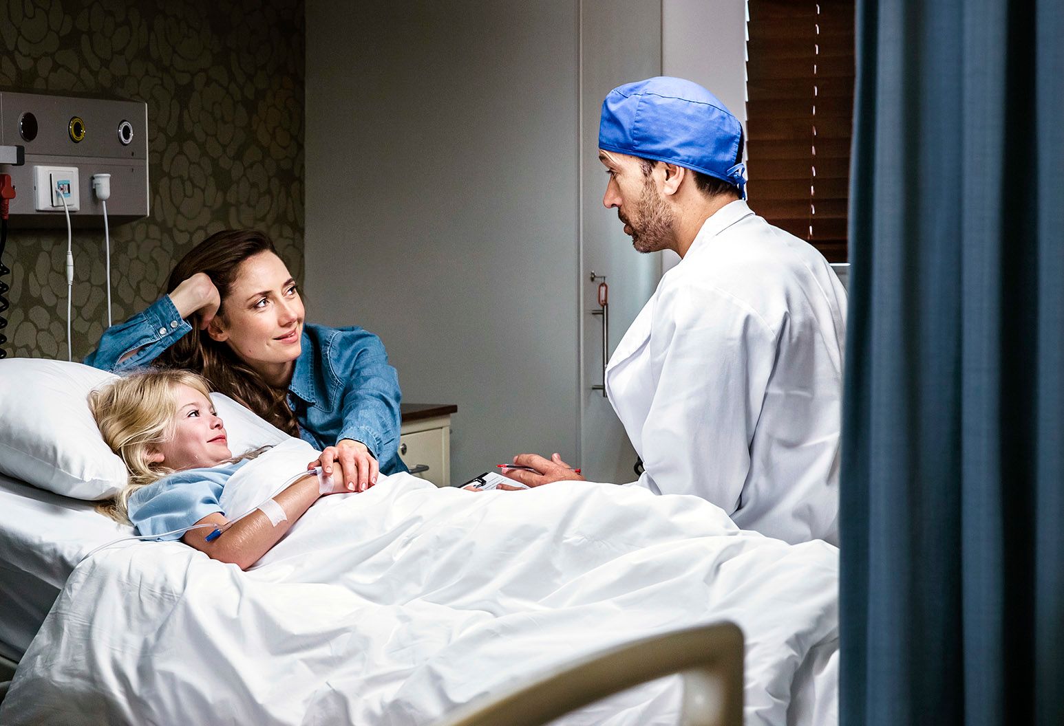 A little girl with blonde hair lays in a hospital bed and her mother is next to her holding her hand. A male doctor wearing a blue operating room cap is also beside, talking to them.