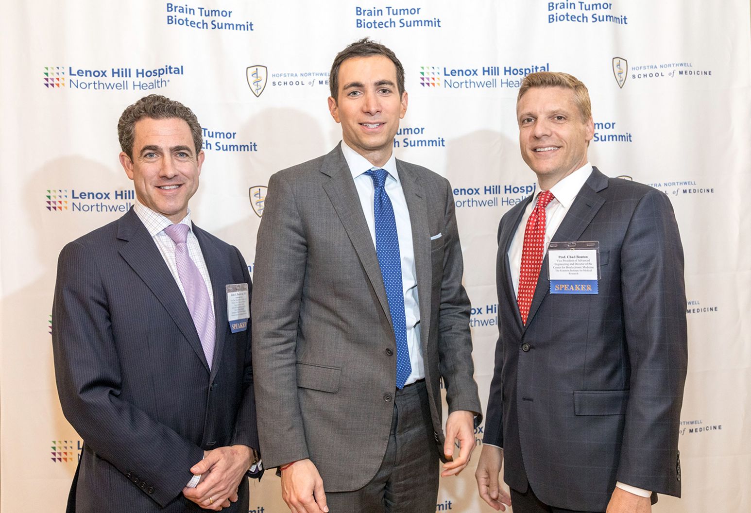 Three male doctors wearing suits are standing in front of backdrop that shows the Lenox Hill Hospital and Hofstra/Northwell School of medicine logos
