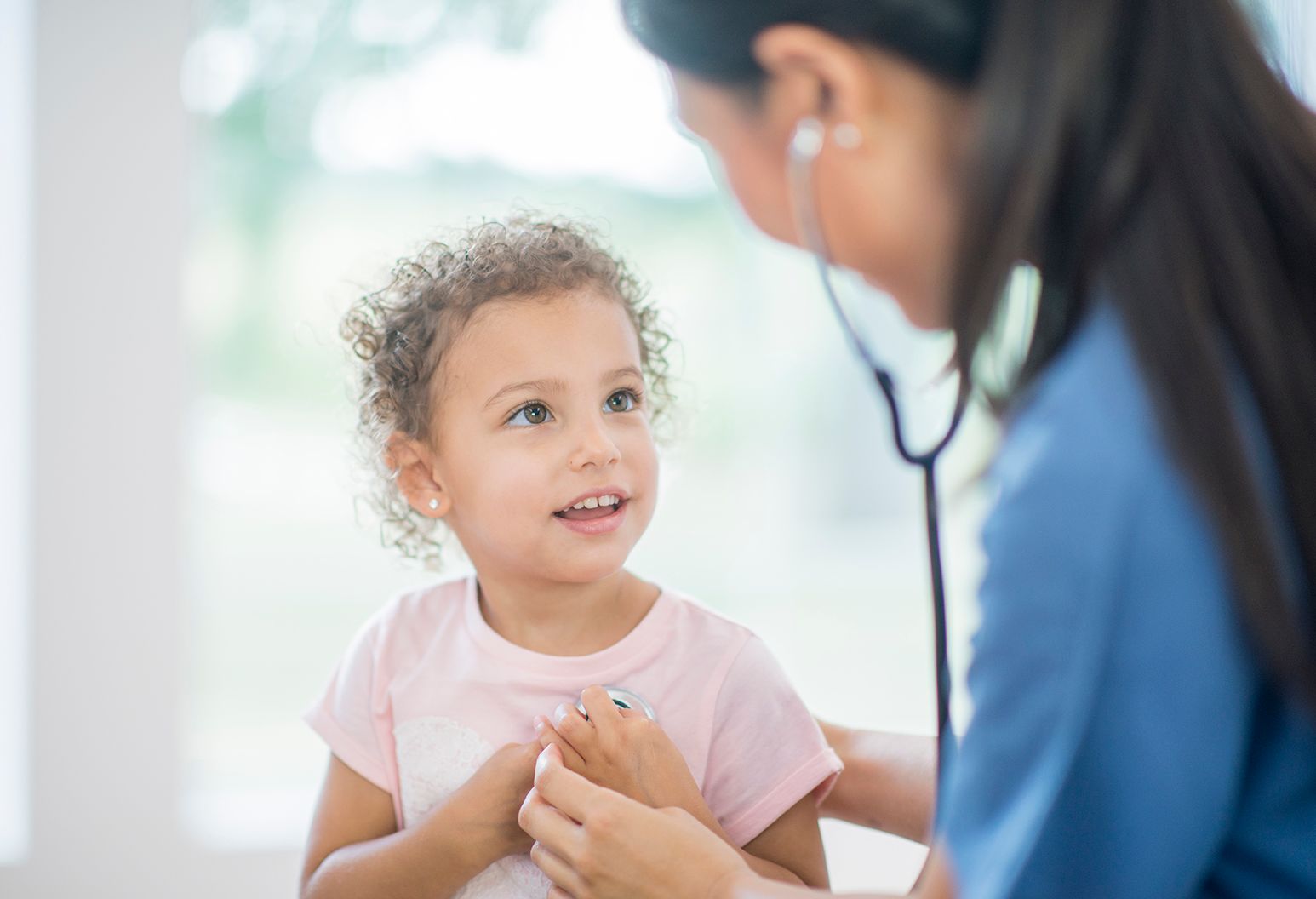 A toddler girl with curly hair is in a doctor's office. She is wearing a pink t-shirt. An Asian female doctor wearing medical clothing is checking her heartbeat with a stethoscope.