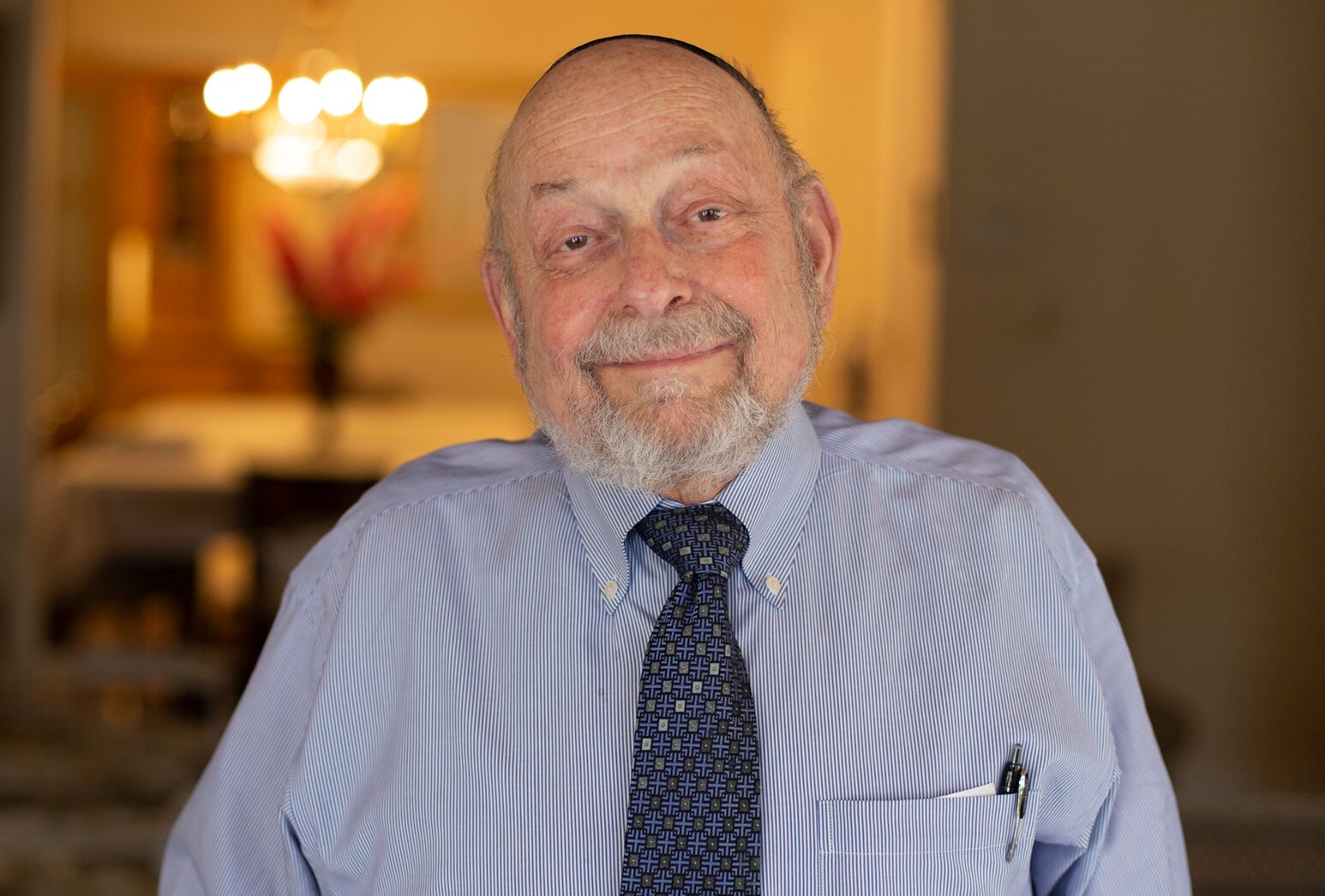 Older man wearing a blue collared shirt and tie smiles in his home.