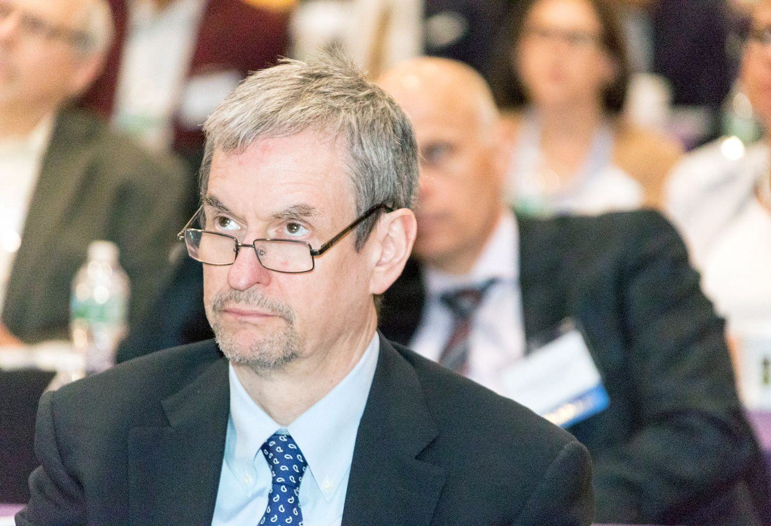 Middle aged man wearing a suit and glasses is watching a presentation at a conference