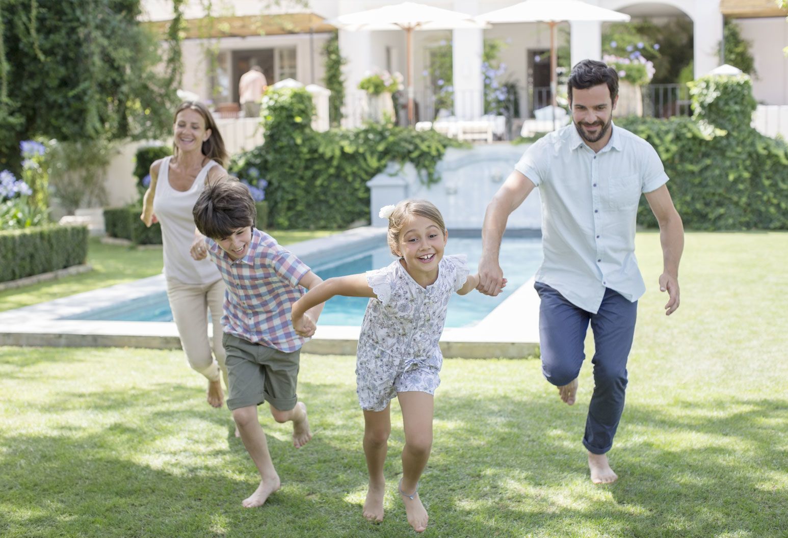 A mom, dad and two young kids hold hands and run together. They are in a backyard with a pool behind them. They're all barefoot and smiling.