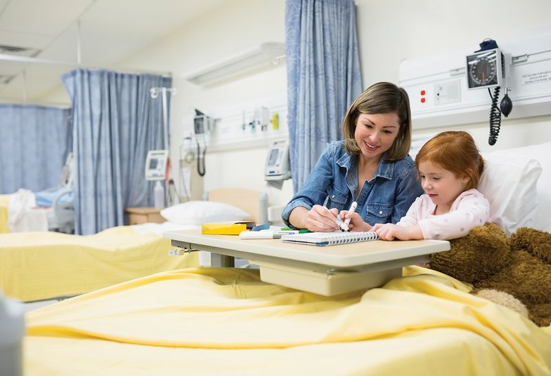 A woman sits next to a four-year-old female pediatric patient and her brown teddy bear in a hospital bed at Cohen Children's Medical Center. Both people are smiling and focused on drawing in a notebook that rests on a table top over the bed. Two empty patient beds with yellow blankets are visible in distance. The blue curtain separators are open.