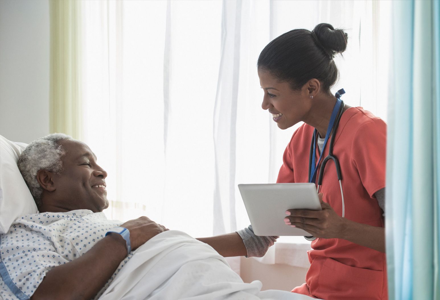An elderly man laying in bed is being looked after by a care professional wearing a stethoscope and holding a tablet.