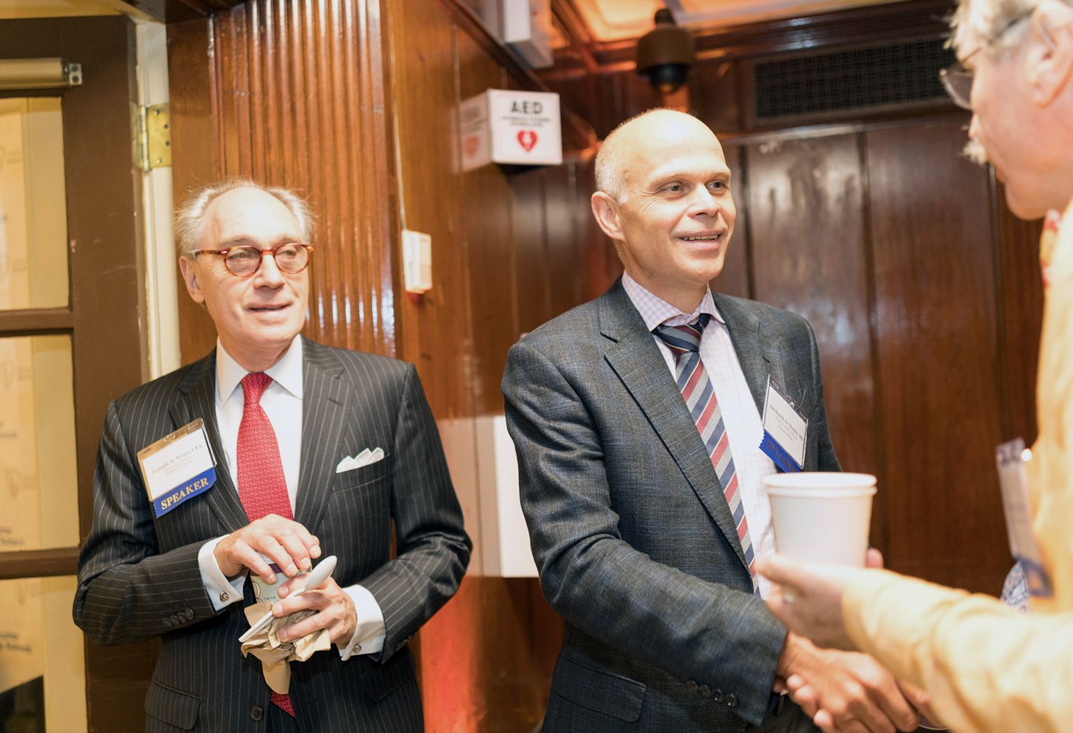 Two older professionals are in a hallway at a conference, talking, while one is shaking the hand of a third man