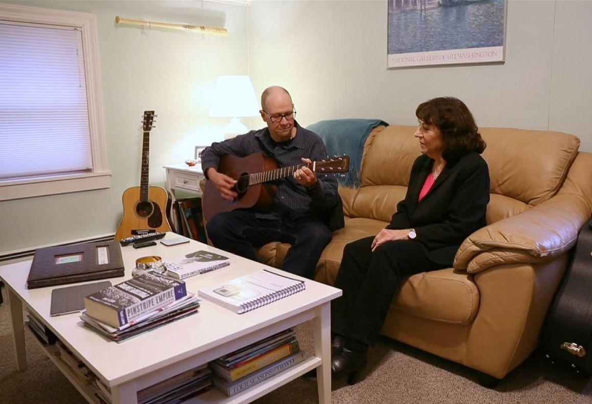 A man sits on a couch playing a guitar. A woman sits next to him and watches on.