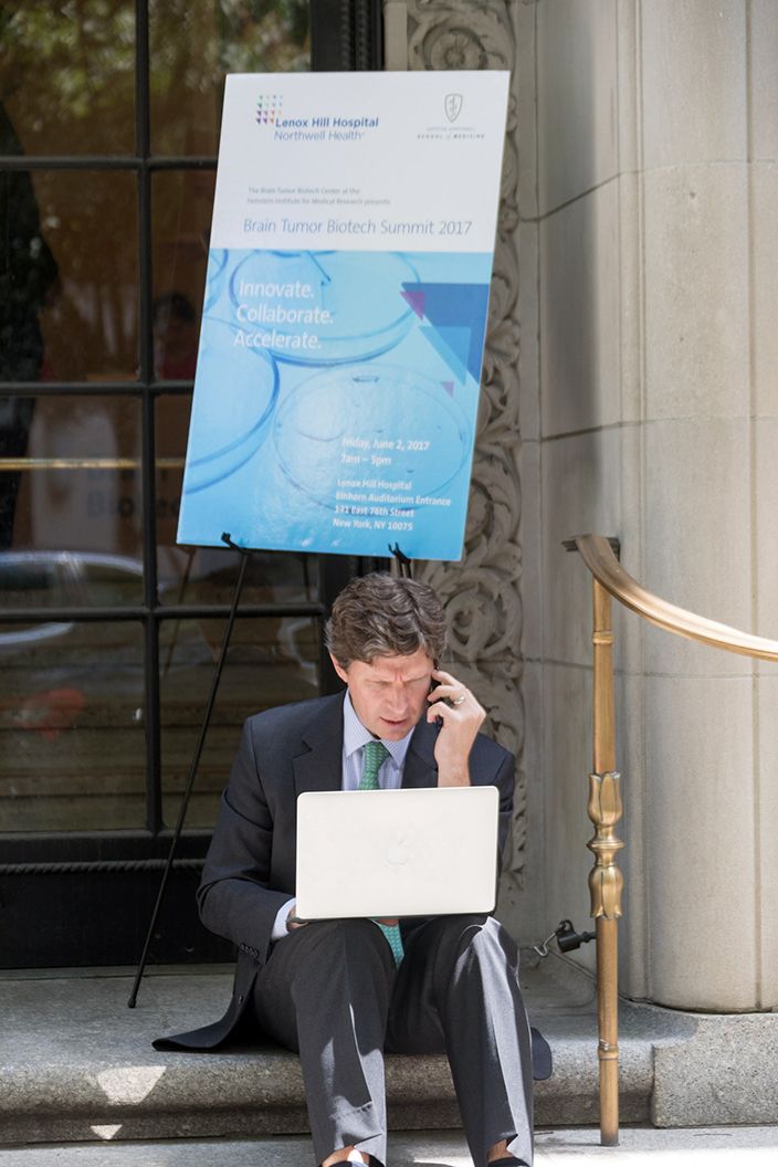 Middle aged man wearing a suit is sitting on the steps outside a building talking on the phone and looking at his computer