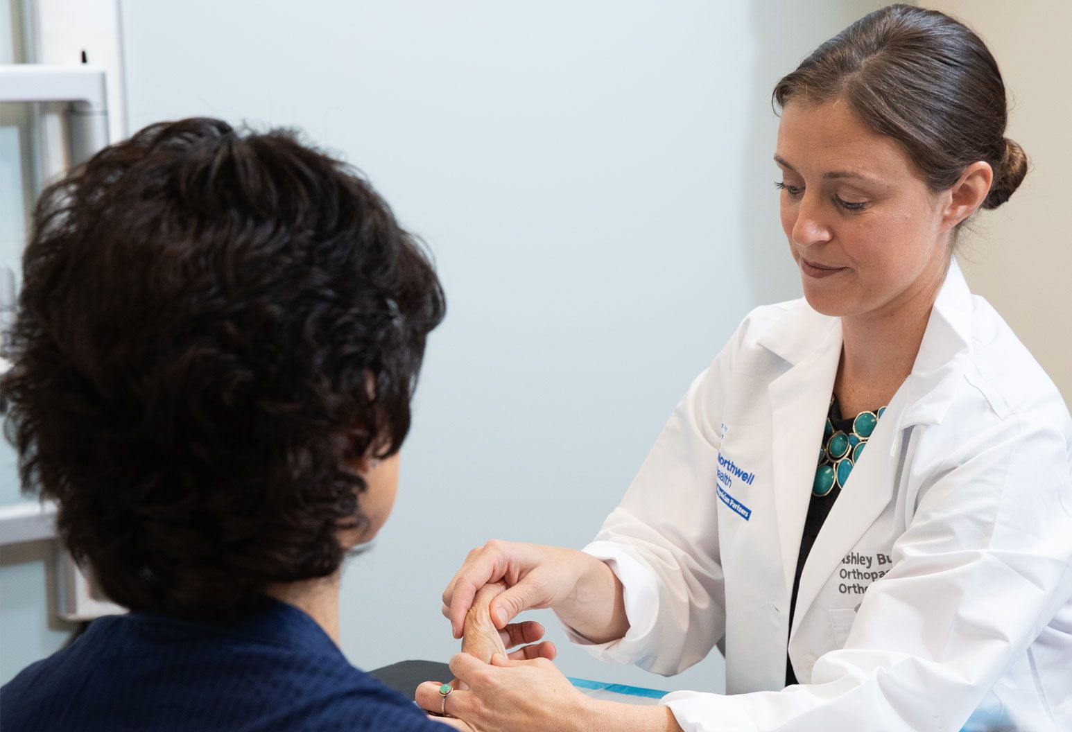 A female doctor examines a female patient's hand. The doctor has brown hair pulled back in a bun and wears a Northwell branded white coat.