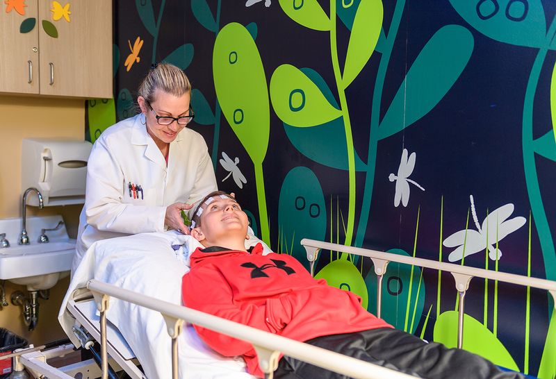 This is a pediatric neurology patient being prepared for an electroencephalogram test in a colorful treatment room. The technician is wearing glasses and adjusting electrodes on the boy's head.