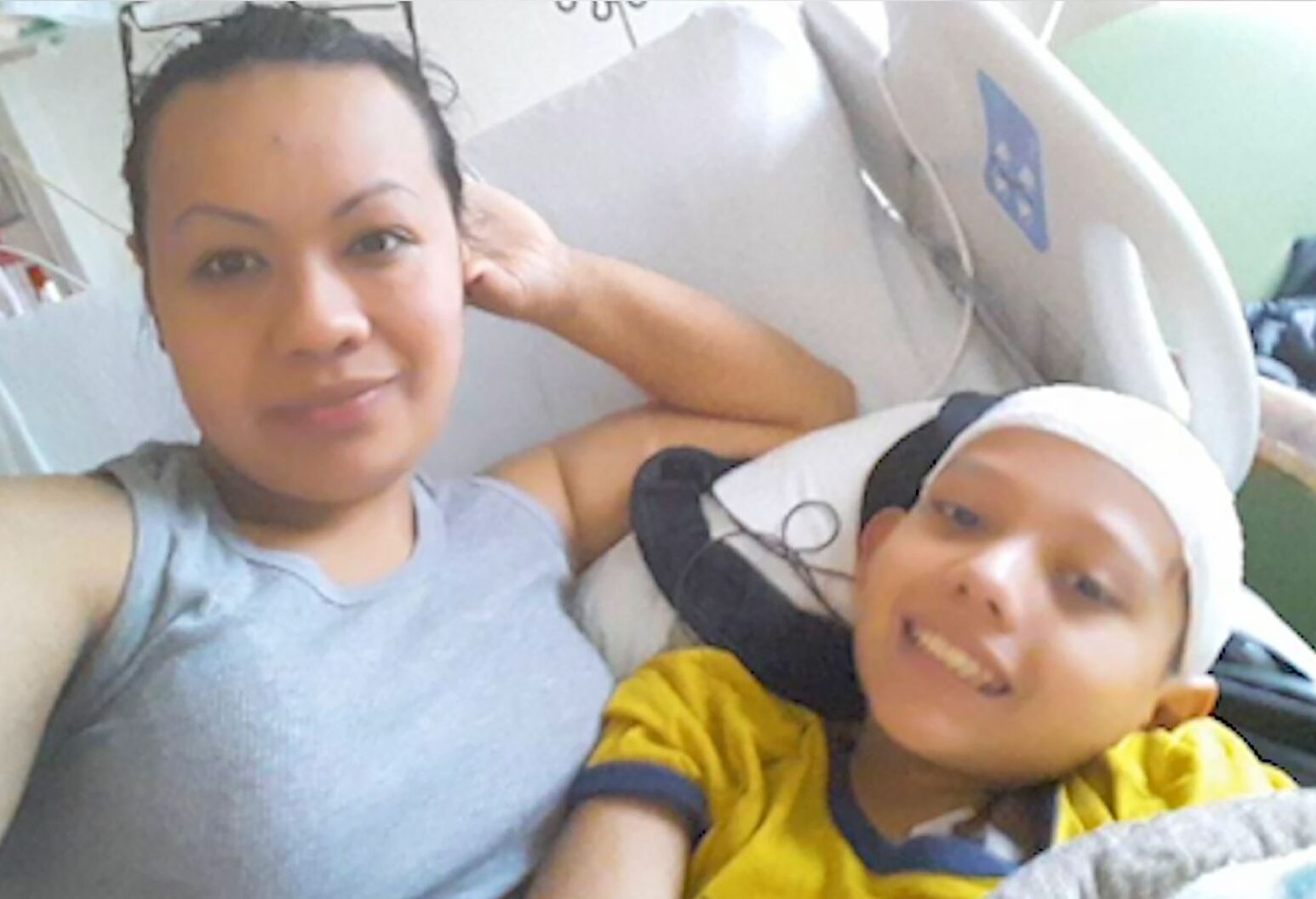 A young boy and his mother lay in a hospital bed, smiling. He had a medical wrap on his head and the mother is wearing a gray tank top.