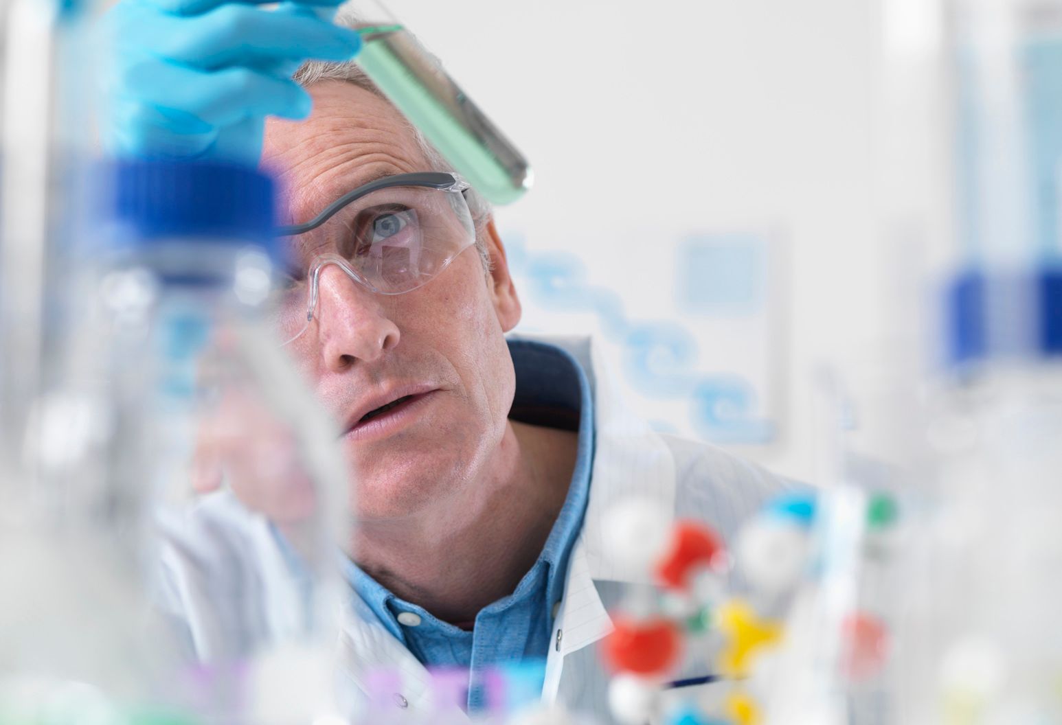 Man wearing a lab coat, gloves and safety glasses, examines a test tube.