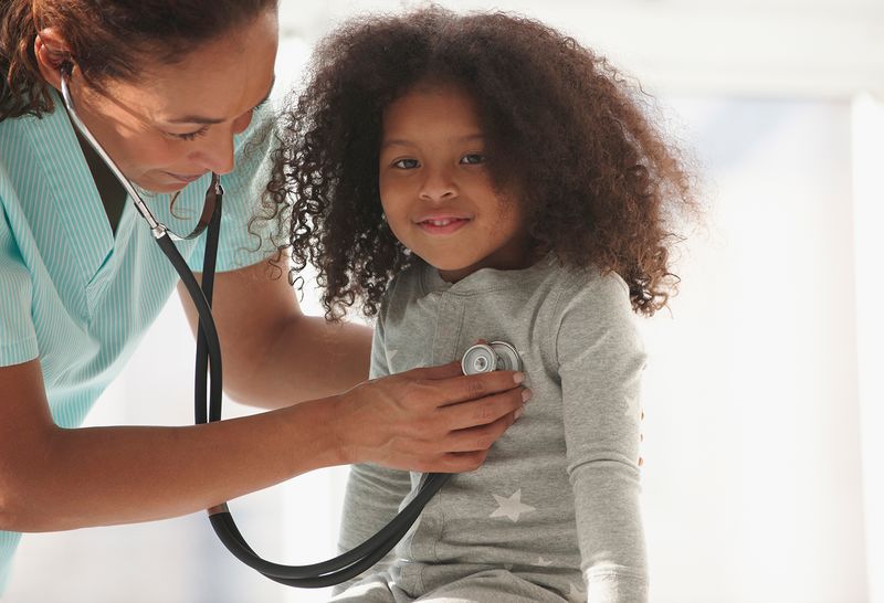 A nurse checks the heart beat of a smiling 5-year-old female pediatric patient using a stethoscope.