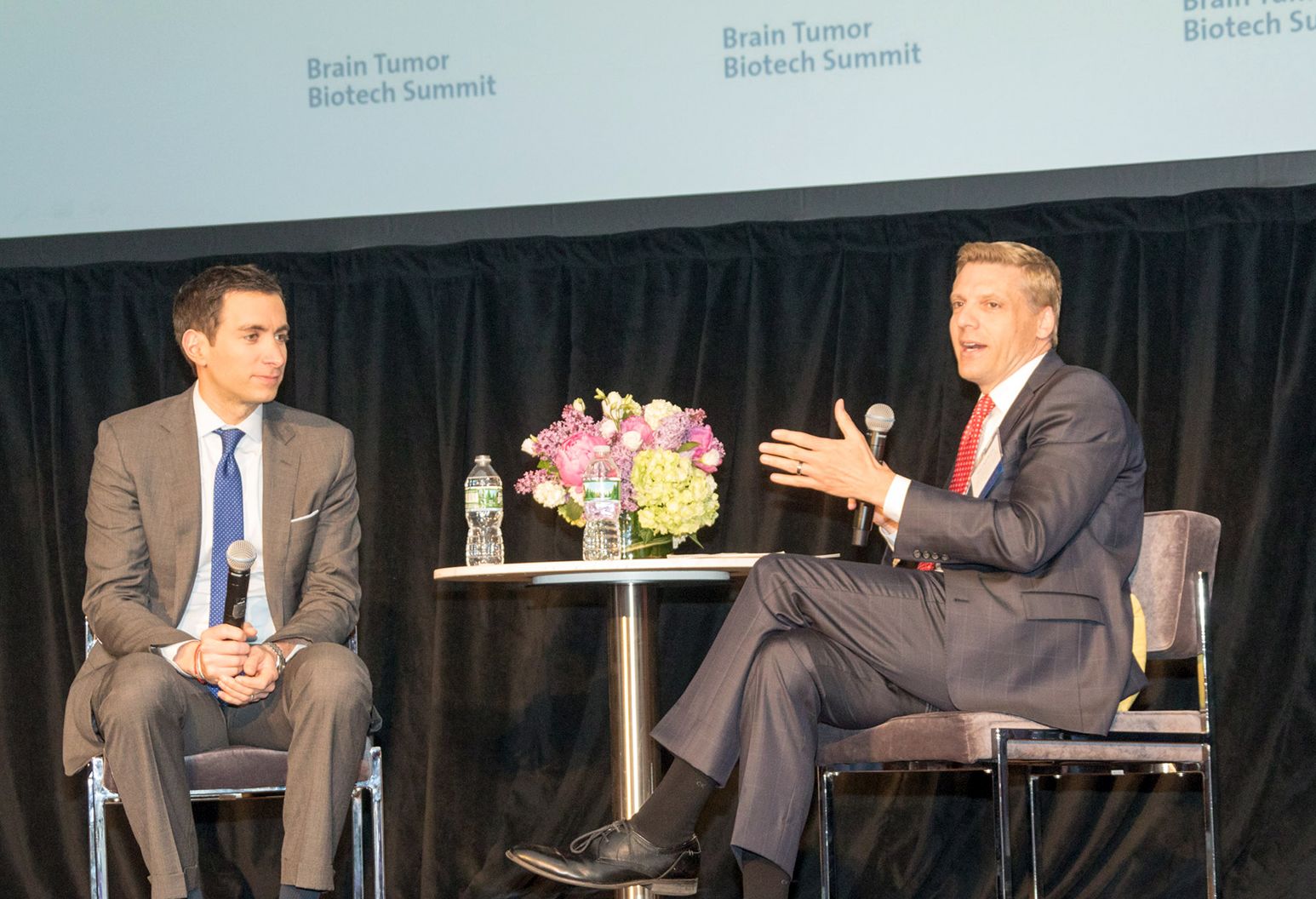 Two men wearing suits are sitting on a stage having a conversation during a conference