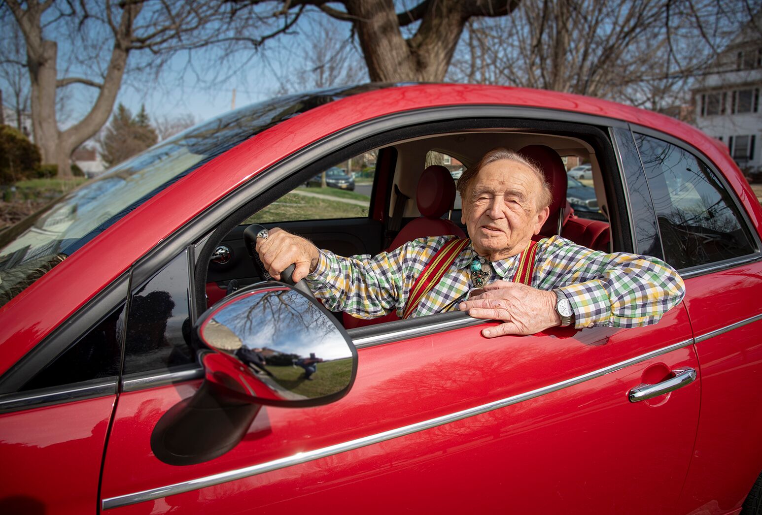 Smiling man in his 90s leaning out of the window of a red car.