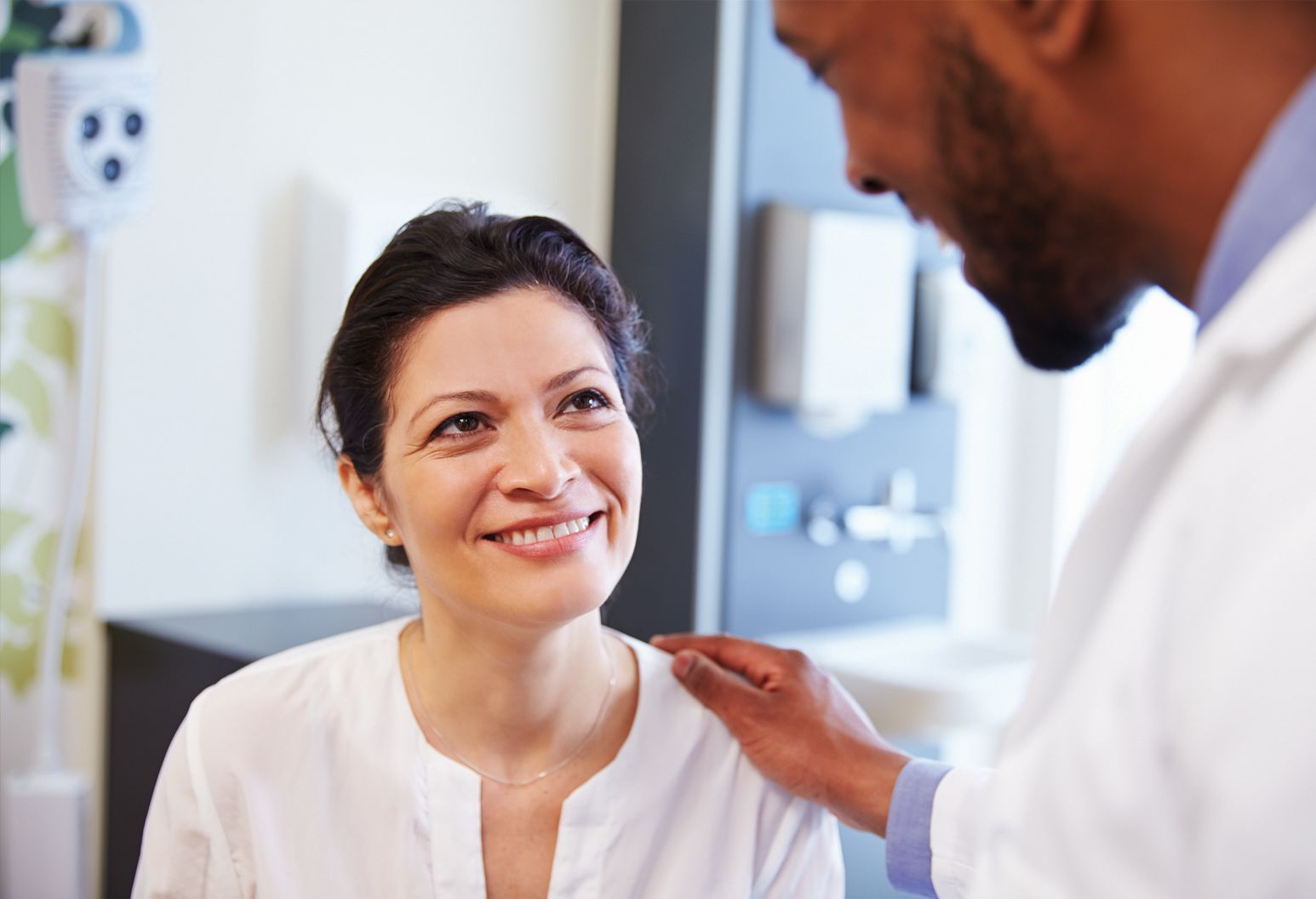 Middle aged woman with brown hair and white blouse smiles as doctor in white lab coat talks to her while gently touching her shoulder.