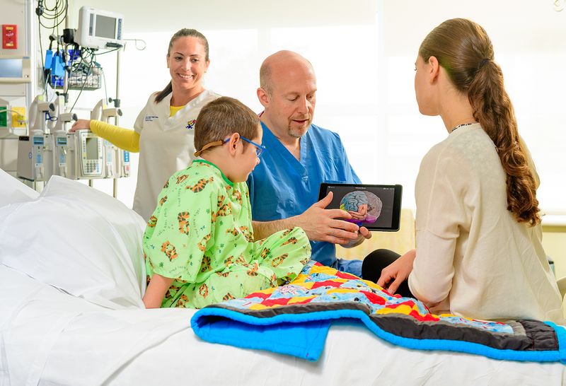 Two medical staff members smile and talk to a 5-year-old male pediatric patient in a bright green hospital gown and his mother, who sits on the hospital bed next to him.