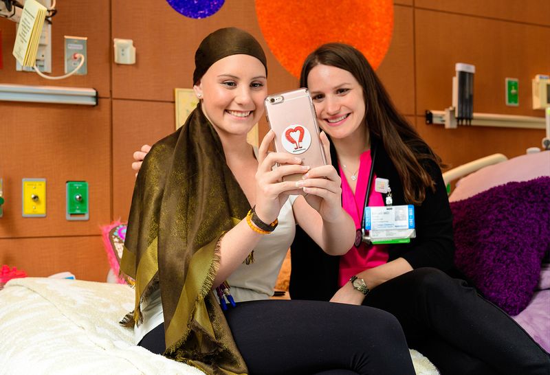 A smiling teenage pediatric patient wears a long brown head scarf. She sits on a Cohen Children's Medical Center hospital bed next to a smiling young female staff member, and the two take a "selfie" together on teen's cell phone.