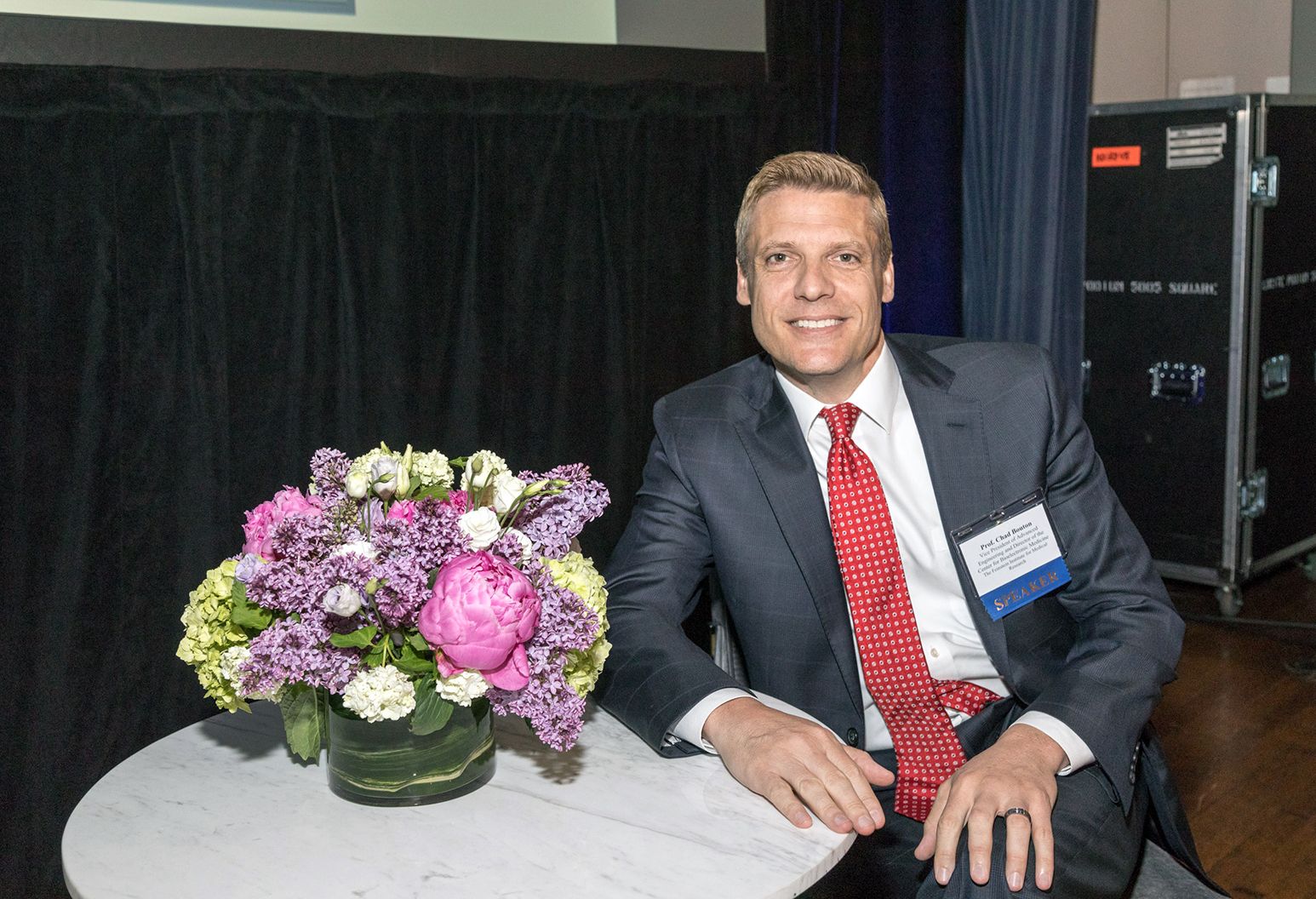 Middle aged man wearing a suit is sitting at a table with a vase of flowers, smiling at the camera