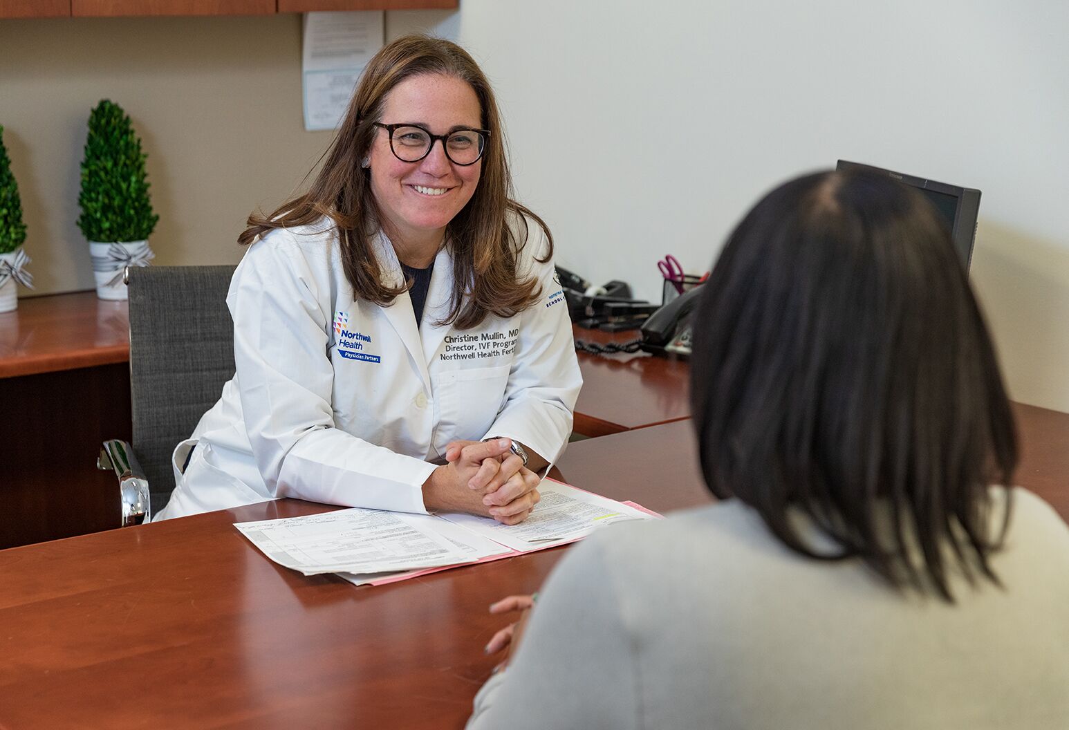 A female doctor wearing a Northwell branded lab coat sits at a desk talking to a female patient.