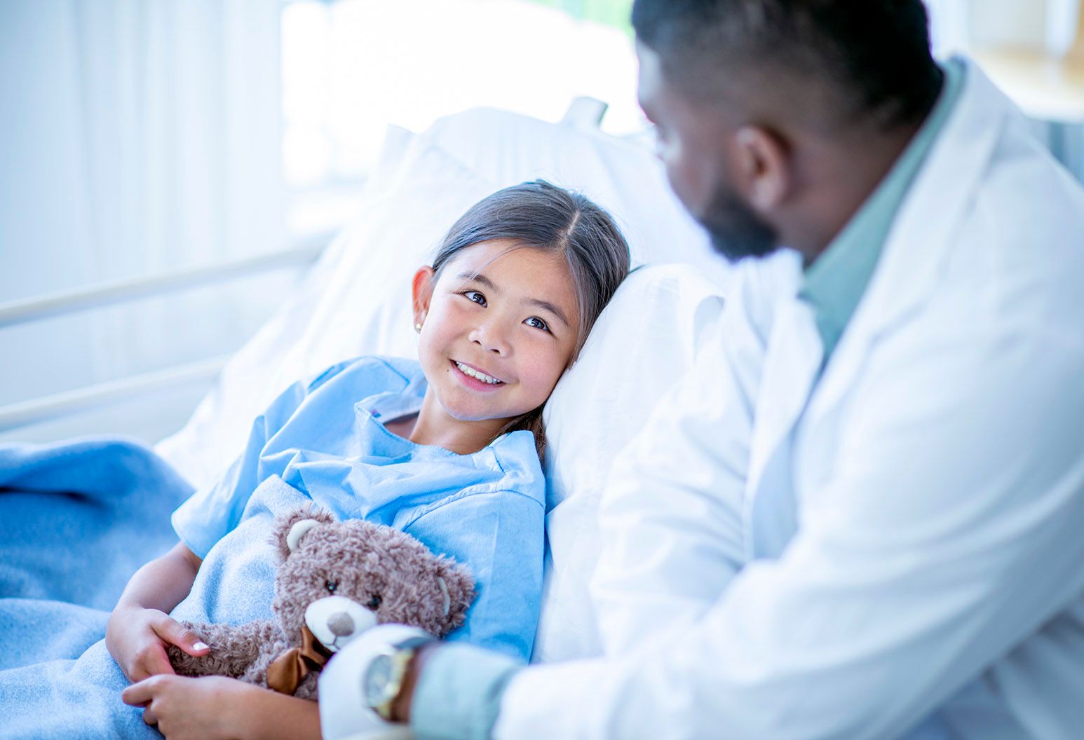 A young girl is laying in bed in a hospital. She is holding a teddy bear while talking with her doctor.