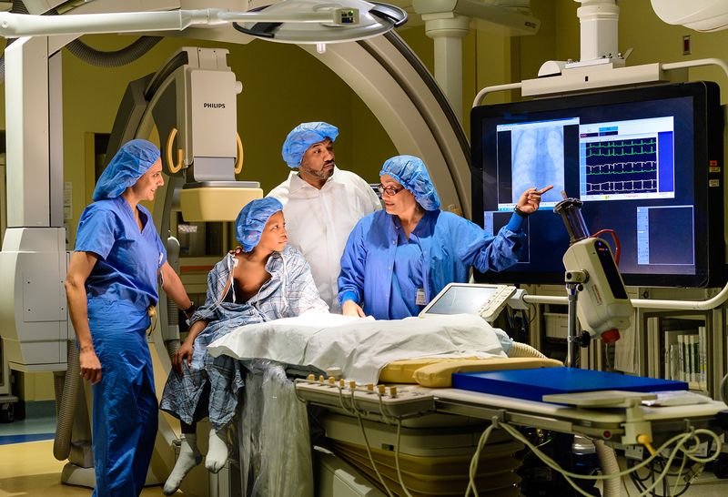 Young patient, around 8 years old, in the operating room with three of our pediatric cardiologists. All are wearing blue surgical caps. The specialists are explaining a procedure to him, pointing at a monitor.