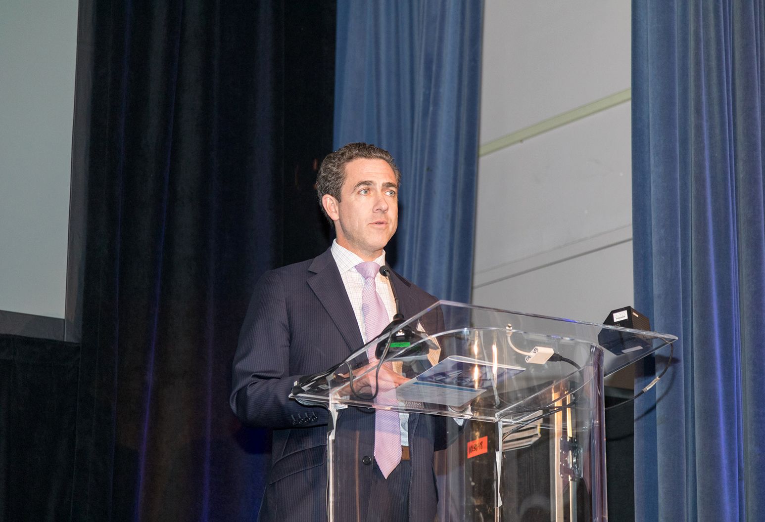 Middle aged man wearing a suit is standing at a podium on stage at a confernece.