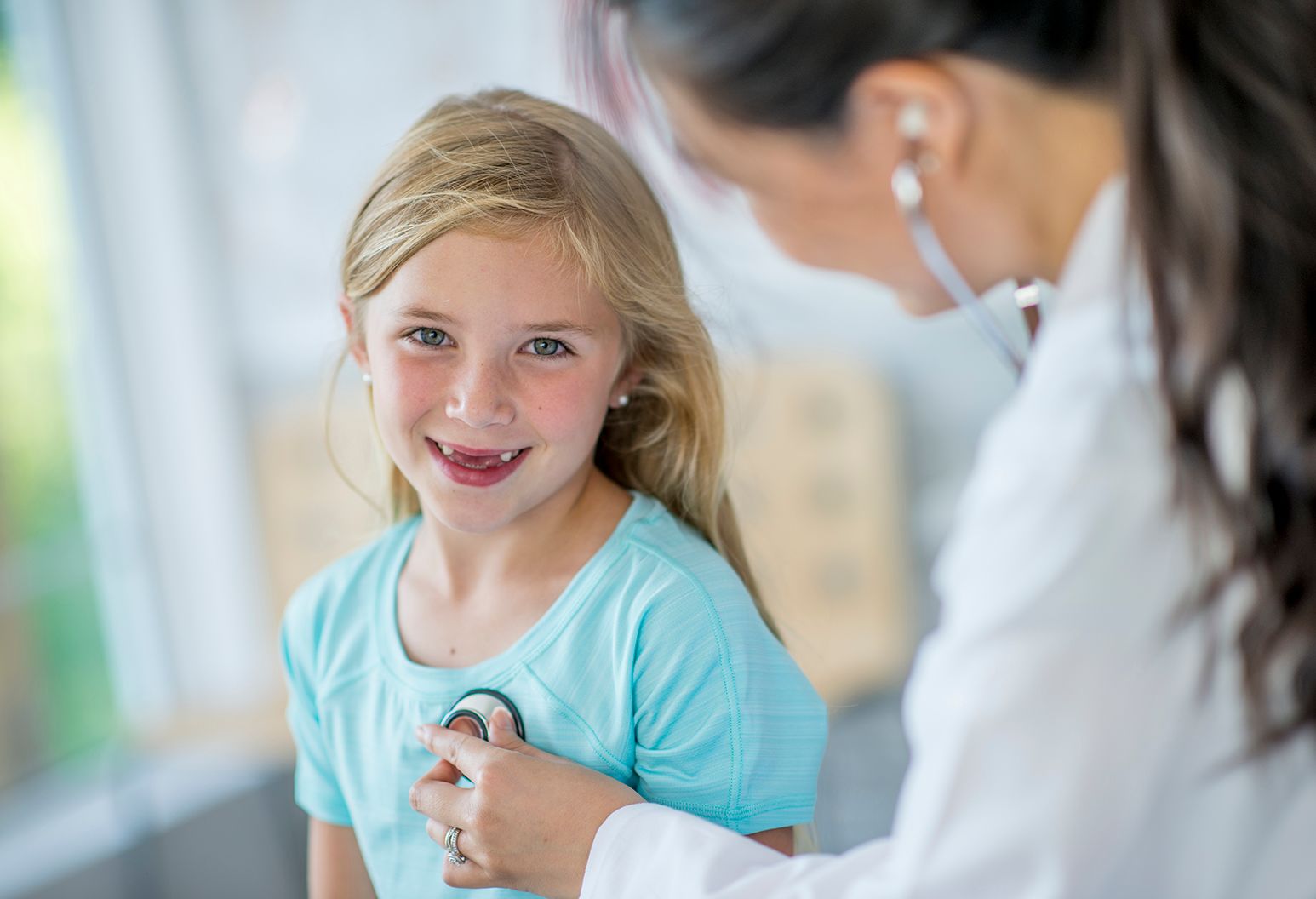 A Caucasian girl and her Asian doctor are indoors in a hospital room. The doctor is checking the girl's heart rate using a stethoscope. The girl is smiling at the camera