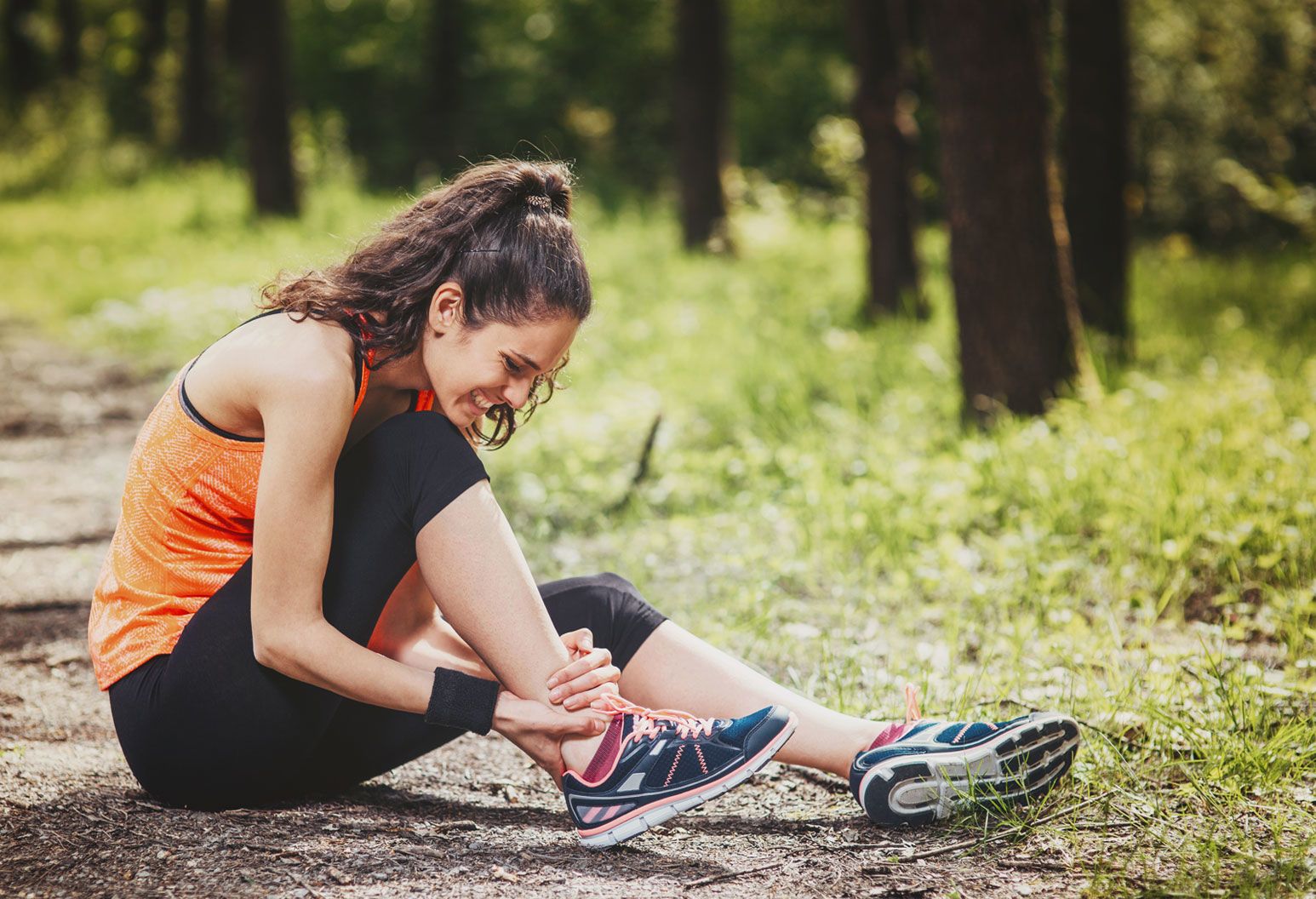 A young woman on a hike sits on the ground and holds her ankle in pain. Her brown hair is in a high pony tail and she's wearing an orange tank top, black leggings and sneakers. She's outside with grass and trees behind her.