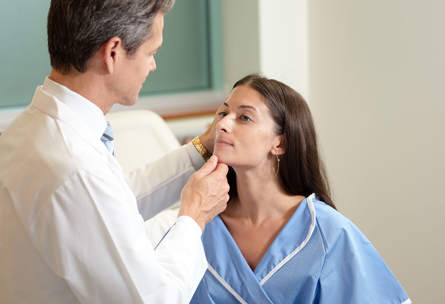 A male doctor in a white lab coat examines the face of a female patient in a blue robe.