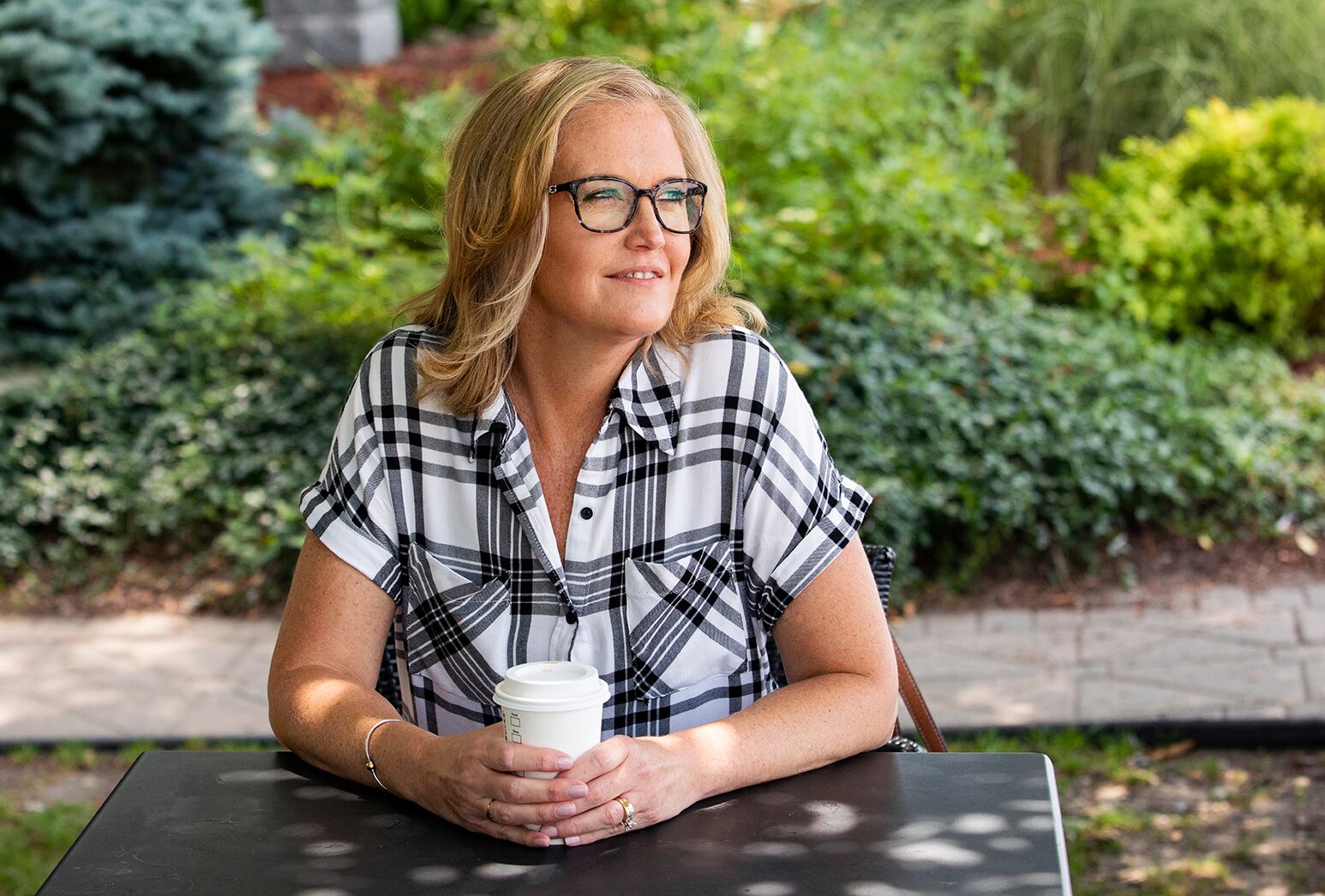Blonde haired woman who had facial plastic surgery in plaid top and glasses, sitting with takeout coffee.