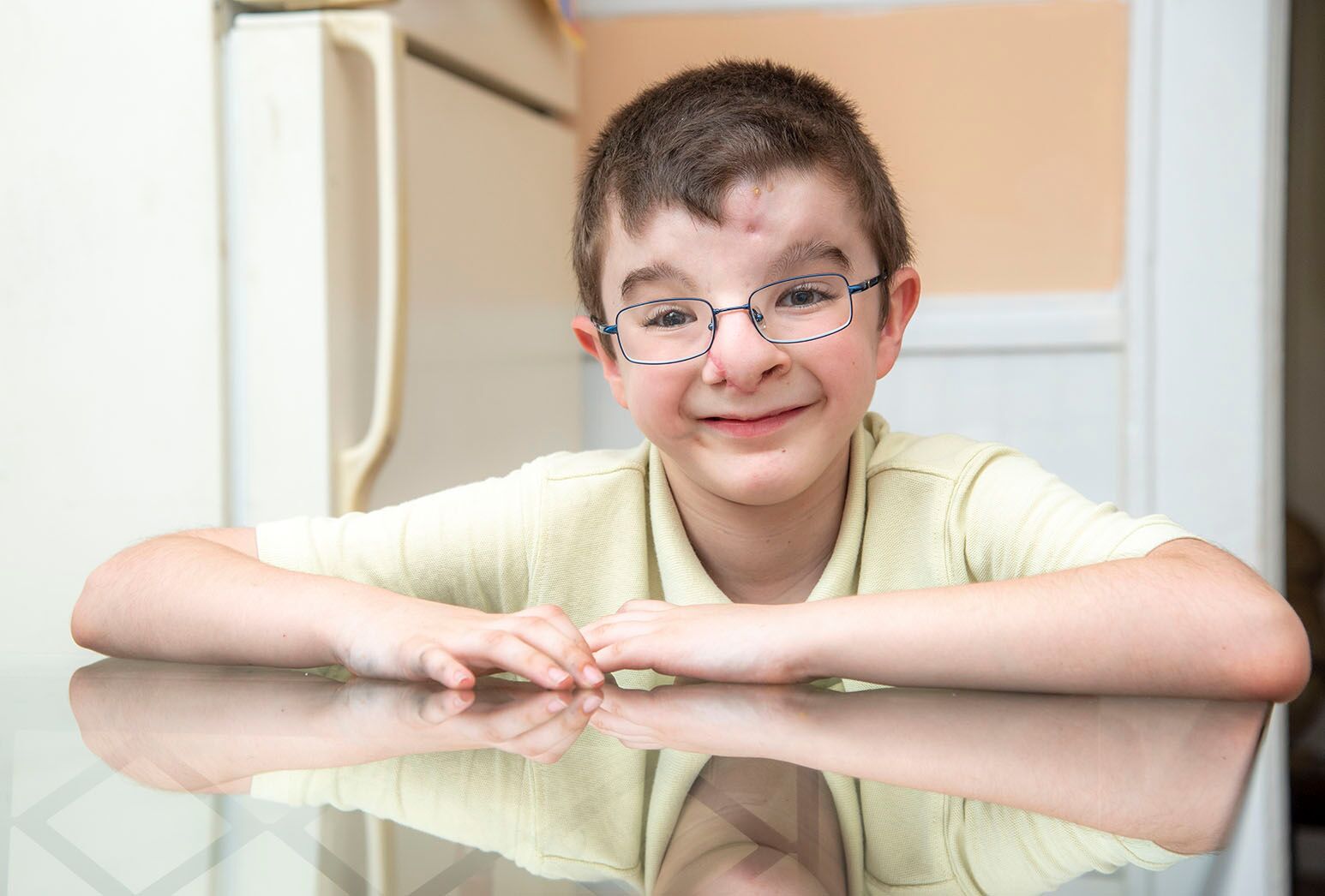 Young boy with dark hair and glasses sits smiling at a glass-topped kitchen table.