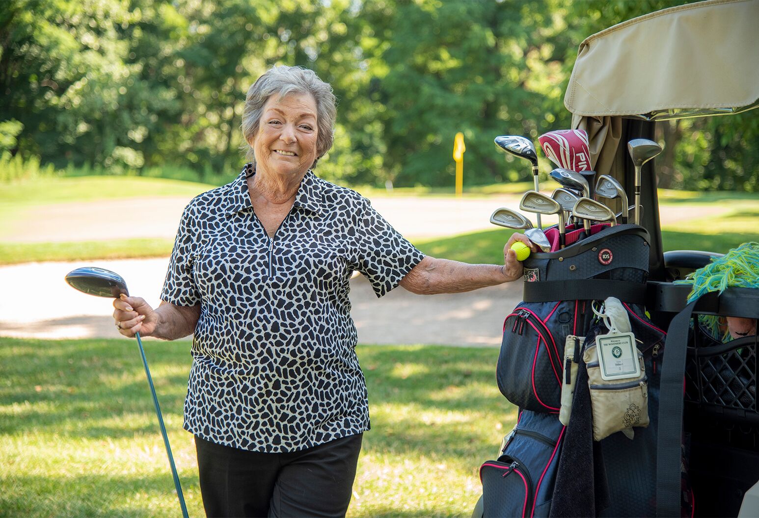Woman in a black and white top and black trousers outdoors with golf clubs and bag.