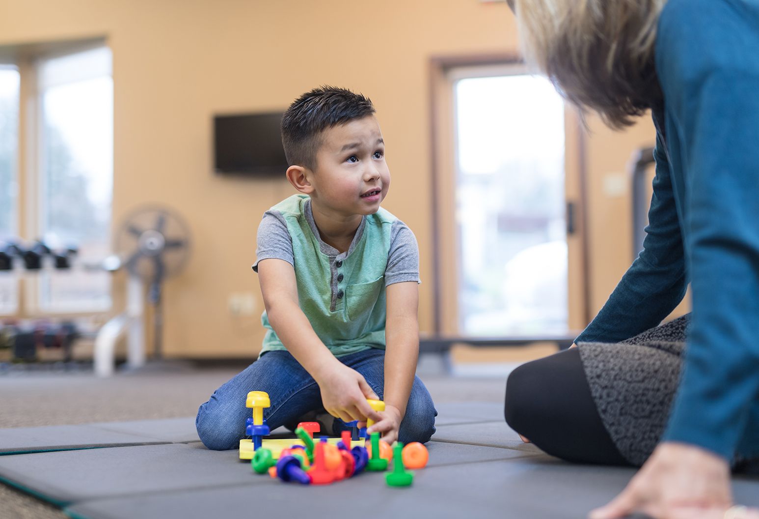 An occupational therapist sits on the floor with an elementary-age boy, working on his fine motor and coordination skills.