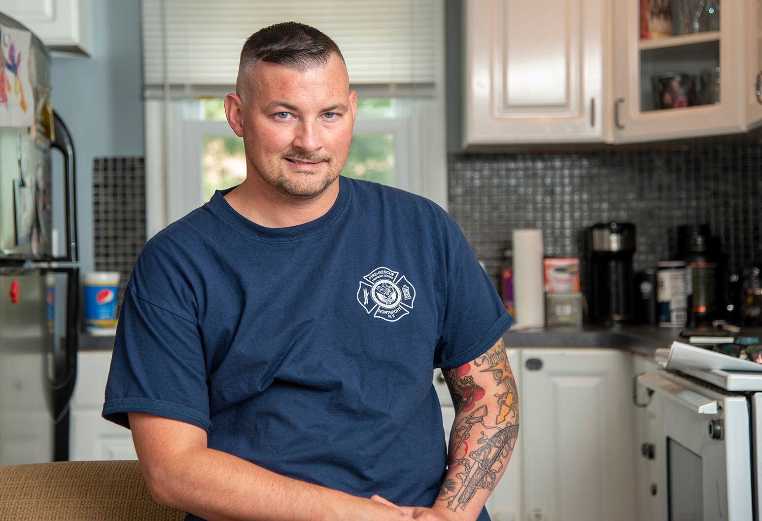 A man with tattoos wearing a navy t-shirt with a firefighter logo smiles in his kitchen.