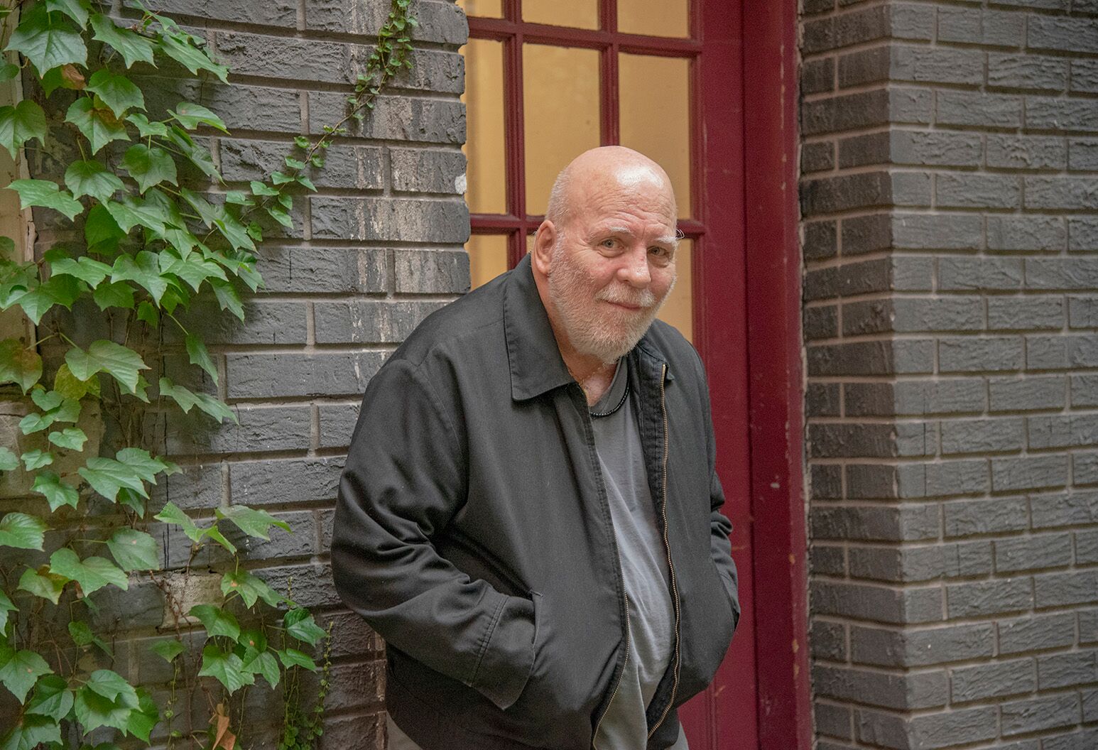Older man in a dark gray coat, leaning against a brick wall and red door with his hands in pockets.
