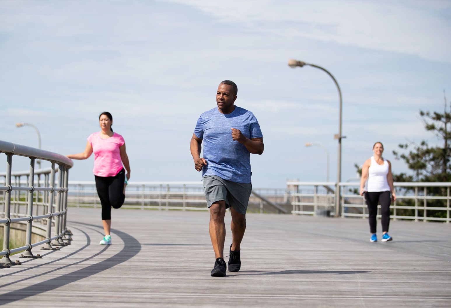 A middle aged man jogs on the beach boardwalk. There are two women out of focus behind him. One is walking and the other is stretching her leg. It's a bright, clear day.
