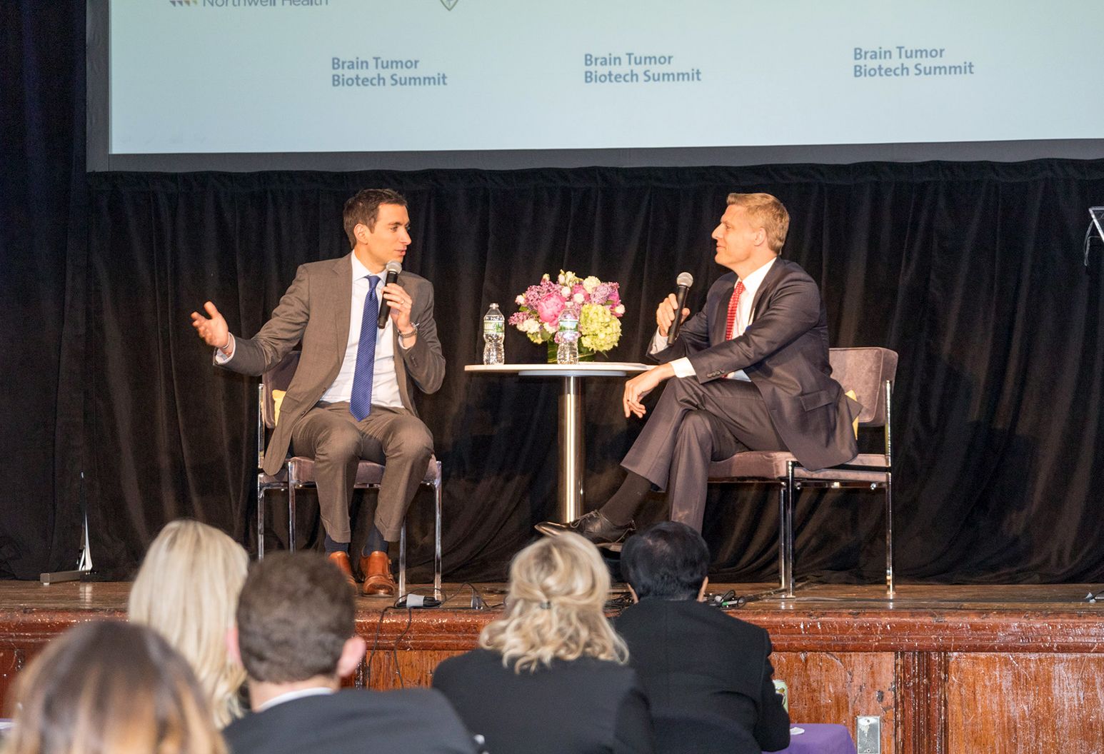 Two men in suites are sitting on a stage having a conversation at a conference