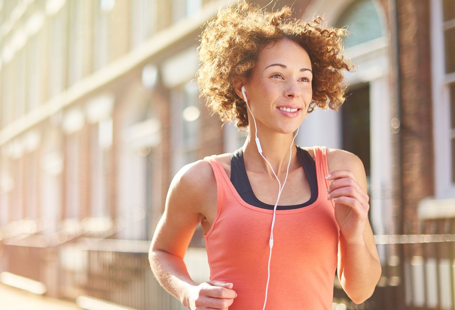A woman jogs outside on a nice day. She wears a bright tank top and headphones.